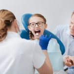 Parental support. Energetic bright teenage lady sitting in the chair while the doctor checking her teeth and the father holding her hand