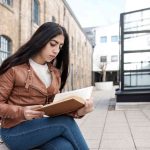 An indian young woman is reading a book sitting on a bench. She is in terrace outdoors and she has very long hair. Casual outfit.